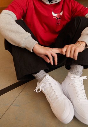 Personne portant un t-shirt rouge des Chicago Bulls, un pantalon noir, des chaussettes grises et des baskets blanches assise en tailleur sur un sol carrelé.