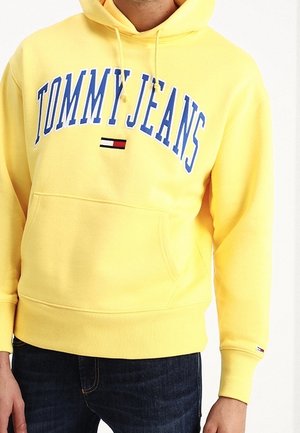 Żółta bluza z kapturem z dużym napisem "TOMMY JEANS" w niebieskich literach oraz małym logo flagi w kolorach czerwonym, białym i czarnym na środku przodu, noszona z ciemnymi jeansami.