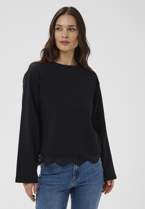 Kvinde med langt mørkt hår iført en sort bluse med lange ærmer og blonder i kanten samt blå jeans, der kigger let til siden.