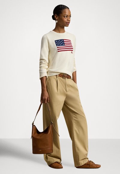 Γυναίκα που φοράει μπεζ πουλόβερ με τη σημαία των ΗΠΑ, μπεζ παντελόνι με φαρδιά ποδόγυρα, καφέ δερμάτινη ζώνη, μοκασίνια και κρατά καφέ δερμάτινη τσάντα χειρός.