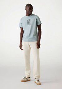 Lichtblauw T-shirt met korte mouwen en een grafische print, gecombineerd met crèmekleurige broek en beige sneakers met groene accenten.