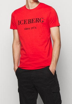 Mężczyzna ubrany w czerwoną koszulkę z krótkim rękawem z napisem "ICEBERG since 1974" oraz czarne spodnie typu cargo, z rękami w kieszeniach, na jednolitym tle.