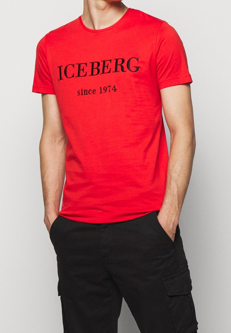 Homme portant un t-shirt rouge à manches courtes avec le texte "ICEBERG depuis 1974" et un pantalon cargo noir, les mains dans les poches, sur un fond uni.