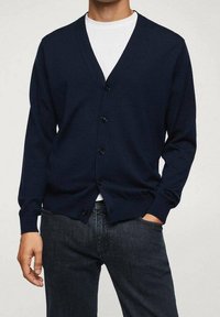 Cardigan bleu marine en tissu texturé, avec un col en V, des manches longues et six boutons sur le devant, porté avec un T-shirt blanc en dessous.