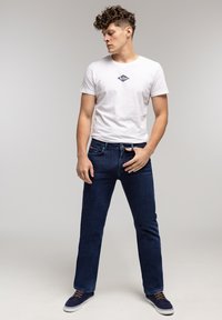 Biała koszulka z ciemnym logo, w połączeniu z granatowymi dżinsami. Casuale trampki z niebieskiego materiału i białą podeszwą. Model stoi w pozycji wyprostowanej.