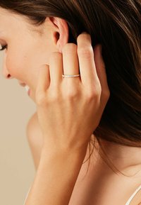 Goldring, besetzt mit kleinen, klaren Steinen, mit einem dünnen Band und einem filigranen Design, getragen an einer Hand, die in das Haar greift. Der Hintergrund ist neutral.