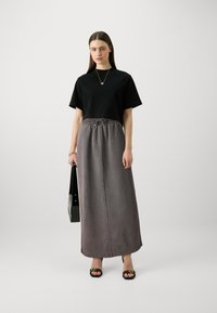 Schwarzes T-Shirt mit kurzen Ärmeln kombiniert mit einem grauen Maxikleid mit elastischem Bund. Das Model hält eine schwarze Handtasche und trägt flache Sandalen.