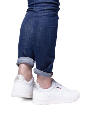 White sneakers with textured material, featuring a logo on the heel, a smooth toe cap, and a rubber sole, worn with cuffed blue jeans.