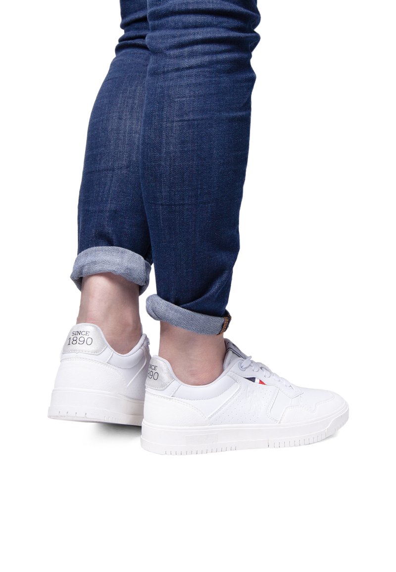 Adidași albi dintr-un material texturat, cu un logo pe toc, o vârf neted și o talpă din cauciuc, purtați cu blugi bleumarin cu manșete.