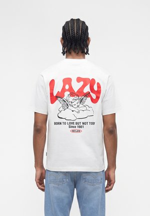 Personne avec des cheveux tressés porte un T-shirt blanc avec le texte rouge "LAZY", illustration d'ange endormi et slogan noir dans le dos.