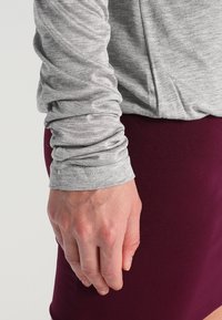 Camicia grigia a maniche lunghe con finitura testurizzata e maniche aderenti, abbinata a una gonna bordeaux. Mano visibile appoggiata vicino al tessuto.