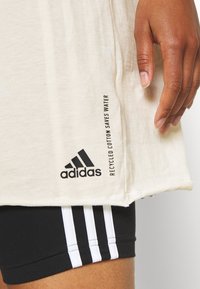 Camiseta beige hecha de algodón reciclado con el logo negro de "adidas" y el texto "EL ALGODÓN RECICLADO AHORRA AGUA" en el lado. Pantalones cortos negros con rayas blancas.