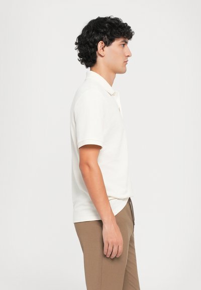 Hvid polo skjorte med korte ærmer og ribbet krave, parret med brune bukser. Enkelt design, lavet af blødt stof, glat tekstur.