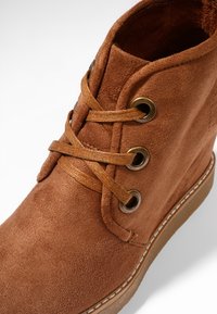 Bottine en daim marron avec trois œillets métalliques et des lacets marron assortis, avec une couture visible le long du bord de la semelle.