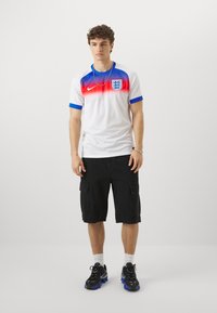Maglia da calcio Nike bianca con spalle a gradiente rosso e blu, con il crest dell'Inghilterra; abbinata a pantaloni cargo neri e sneakers.