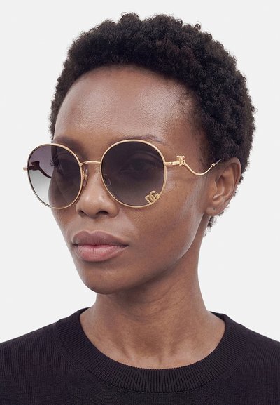 Persona con capelli corti e ricci che indossa grandi occhiali da sole rotondi sfumati con montature dorate e una maglietta nera su uno sfondo semplice.