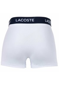 Fehér pamut boxer alsónadrág sötétkék derékvonallal, amelyen a "LACOSTE" logó látható. Simább textúra, közepes magasságú dizájn, minimális varrási részletekkel.