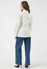Witte katoenen blouse met lange mouwen en een tailleband, gecombineerd met losse blauwe denim broek en puntige zwarte hakken. Eenvoudig en strak ontwerp.