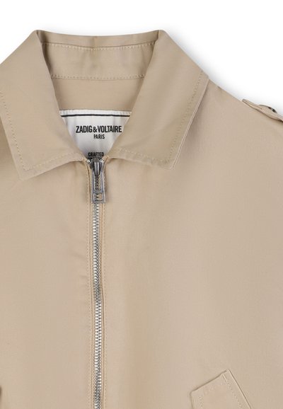 Veste légère beige avec un col pointu, une fermeture éclair à l'avant et une poche plaquée, en tissu lisse à texture minimale.