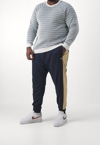 Tröja med ljusblå och vita horisontella ränder, ribbade mudd. Mörkblå träningsbyxor med beige sidopanel. Vita sneakers med bruna accenter.