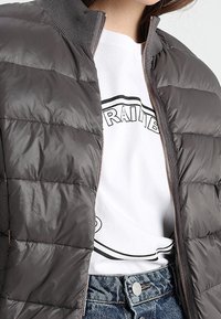 Chaqueta acolchada gris oscuro con cierre frontal, tejido texturizado y cuello de canalé, usada sobre una camiseta gráfica blanca y vaqueros azules.