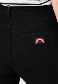 Nahaufnahme der hinteren Tasche einer schwarzen Jeans mit rotem und weißem Wrangler-Logo-Patch am äußeren Taschenrand.