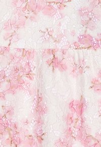 Krajková tkanina s květinovým vzorem v jemné růžové a bílé, s propracovanými texturami a jemnými hnědými akcenty. Poloprůhledný design.