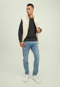 Beige bolyhos mellény sötét, texturált pulóver felett, világoskék farmernadrággal és fehér sportcipővel párosítva. A modellen enyhe szögben áll.