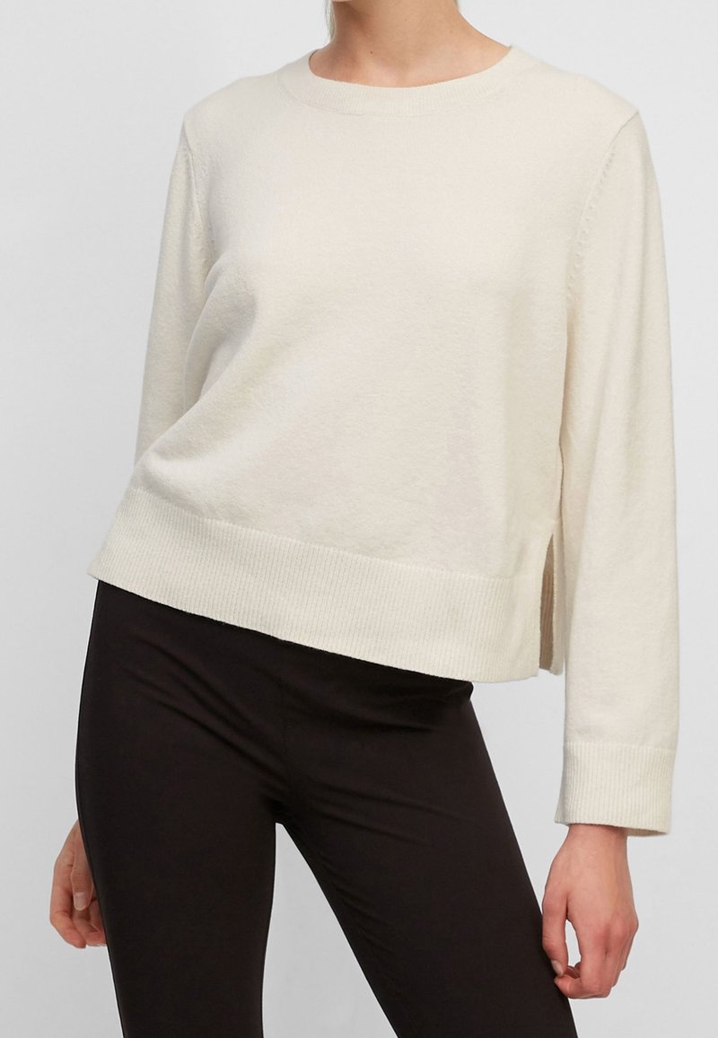 Femme portant un pull à manches longues de couleur crème avec des fentes latérales et un pantalon noir ajusté, debout devant un fond uni.