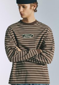 Barna hosszú ujjú póló fehér vízszintes csíkokkal és központi ovális Ford logóval. Puha anyagból készült, laza szabással.