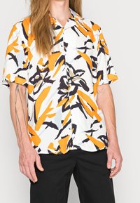 Short-sleeve shirt in white with bold orange, black, and gray abstract patterns. Lightweight fabric with a relaxed fit and pointed collar.