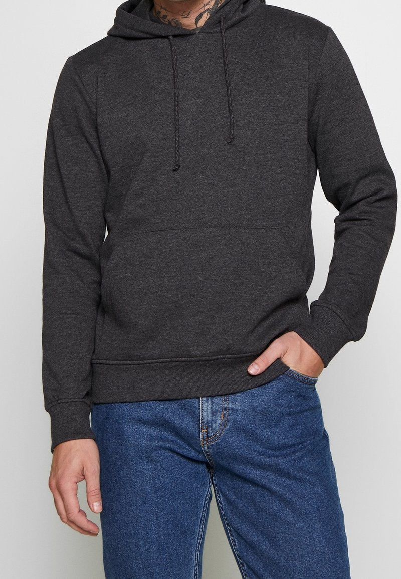 Homme portant un sweat à capuche gris foncé et un jean bleu, une main dans la poche, sur fond uni.
