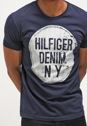 Mężczyzna ubrany w granatowy t-shirt z dużym białym okrągłym nadrukiem z napisem "HILFIGER DENIM N Y" na przodzie.