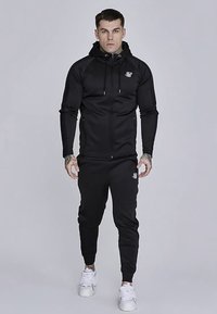 Črna jakna z zadrgo in kapuco, oprijetega kroja, z logotipom na prsih, kombinirana s usklajenimi trenirkami in belimi supergami. Gladek teksturni vzorec tkanine.