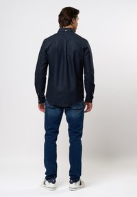 Marineblaues Hemd mit langen Ärmeln, Rückenpasse, taillierter Schnitt, kombiniert mit blauen Jeans und weißen Turnschuhen.