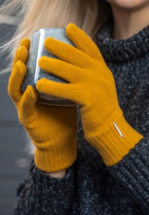 Guantes de lana amarillos con puños acanalados, usados mientras se sostiene una taza de cerámica azul, sobre un fondo de suéter oscuro y texturizado.