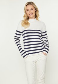 Pulover tricotat cu nervuri, de culoare crem, cu dungi orizontale bleumarin. Guler înalt, mâneci lungi cu manșete late, asortat cu pantaloni albi.