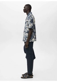 Cămașă cu mânecă scurtă de culoare albastră închisă, cu un model floral alb, asortată cu pantaloni întunecați și sandale negre. Materialele și texturile nu sunt specificate.