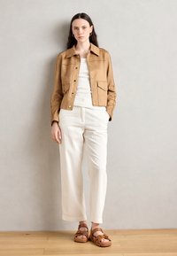 Giacca scamosciata beige con bottoni a pressione, abbinata a una maglietta bianca e pantaloni in maglia color crema. Sandali marroni completano l'outfit. Design semplice e testurizzato.