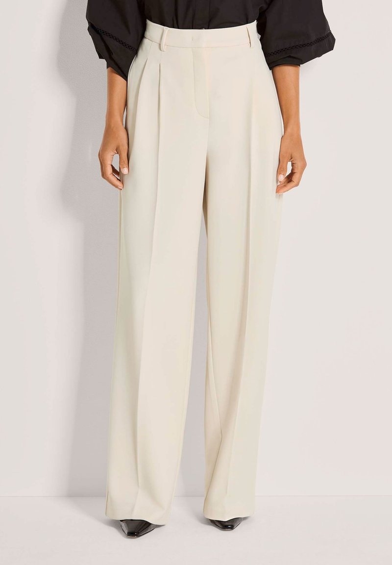 Cremefarbene, weit geschnittene Hosen mit maßgeschneidertem Design, vorne mit Falten und glatter Textur. Kombiniert mit schwarzen spitzen Pumps.
