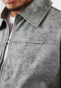 Grijze zip-up jas met een gestructureerde afwerking, grote kraag en subtiel gemêleerd patroon. Bevat een klein metalen accent op de borst.