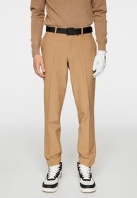 Beige skräddarsydda byxor med rak benpassform, tillverkade av lättviktsmaterial. Svart perforerat bälte, kombinerat med svarta och vita sneakers.