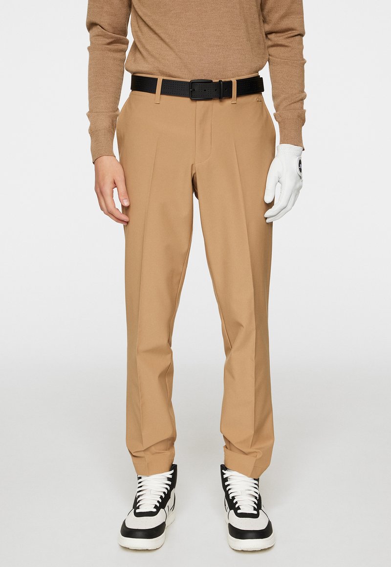 Beige skräddarsydda byxor med rak benpassform, tillverkade av lättviktsmaterial. Svart perforerat bälte, kombinerat med svarta och vita sneakers.