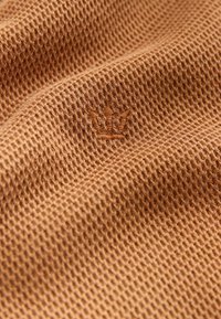 Tejido de punto en waffle marrón con textura y un logo de corona bordado. La trama es suelta, proporcionando una apariencia transpirable y ligera.