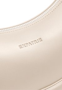 Beige Tasche aus synthetischem Leder mit glatter Textur, geschwungenem Design und eingeprägtem Logo, mit einem silbernen Reißverschluss entlang der oberen Kante.