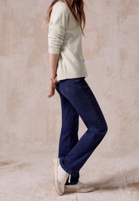 Žena stojící bokem, oblečená v béžovém svetr, tmavě modrých džínách a bílých teniskách, na béžovém strukturovaném pozadí.