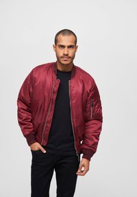 Veste bomber bordeaux avec fermeture éclair à l'avant, poignets côtelés et poche sur le côté. Portée par-dessus une chemise noire avec une texture lisse et brillante.