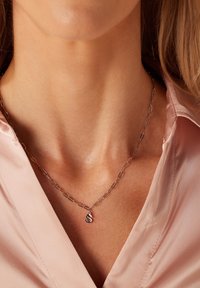 Zilveren ketting met verlengde schakels en een klein hanger met de letter "S," gedragen over een lichtroze satijnen shirt.