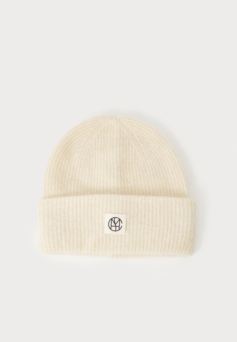 Knit cream beanie with ribbed texture, featuring a fold-over cuff and a small black logo patch. Soft material and rounded shape.