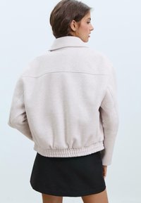 Veste en polaire rose clair avec col haut, ourlet élastique et détails de couture aux épaules, associée à une jupe noire. Vue de dos.